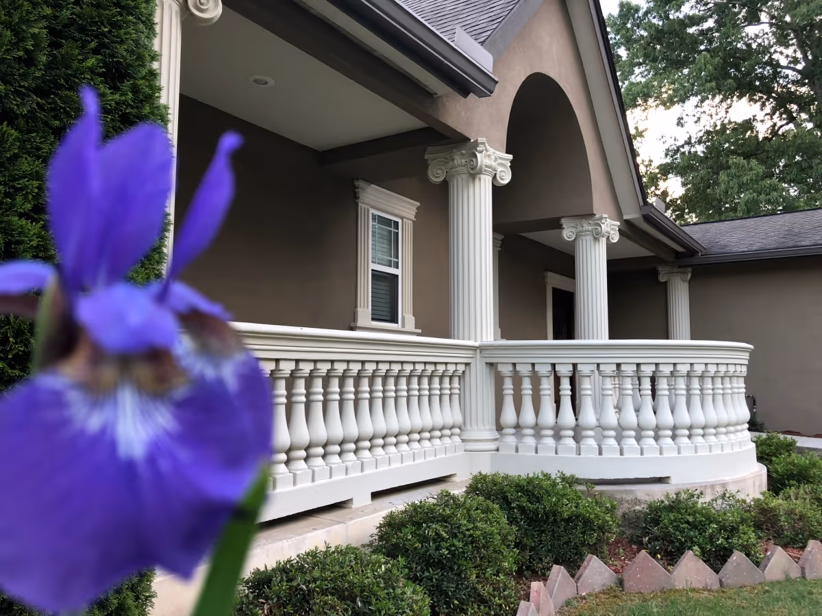 Close-up of a purple flower in the foreground with a beige building featuring white classical columns and a white balustrade in the background, surrounded by green bushes and trees.