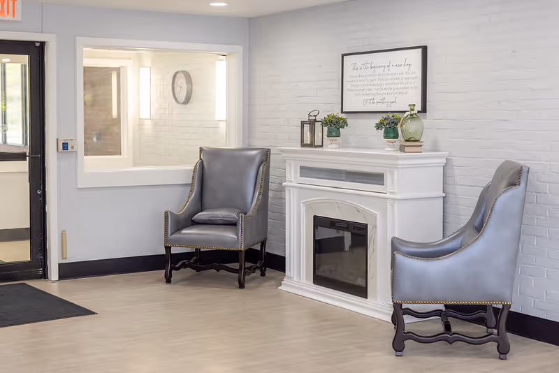 Two gray armchairs flank a white electric fireplace mantel in a bright seating area with framed art and small plants.