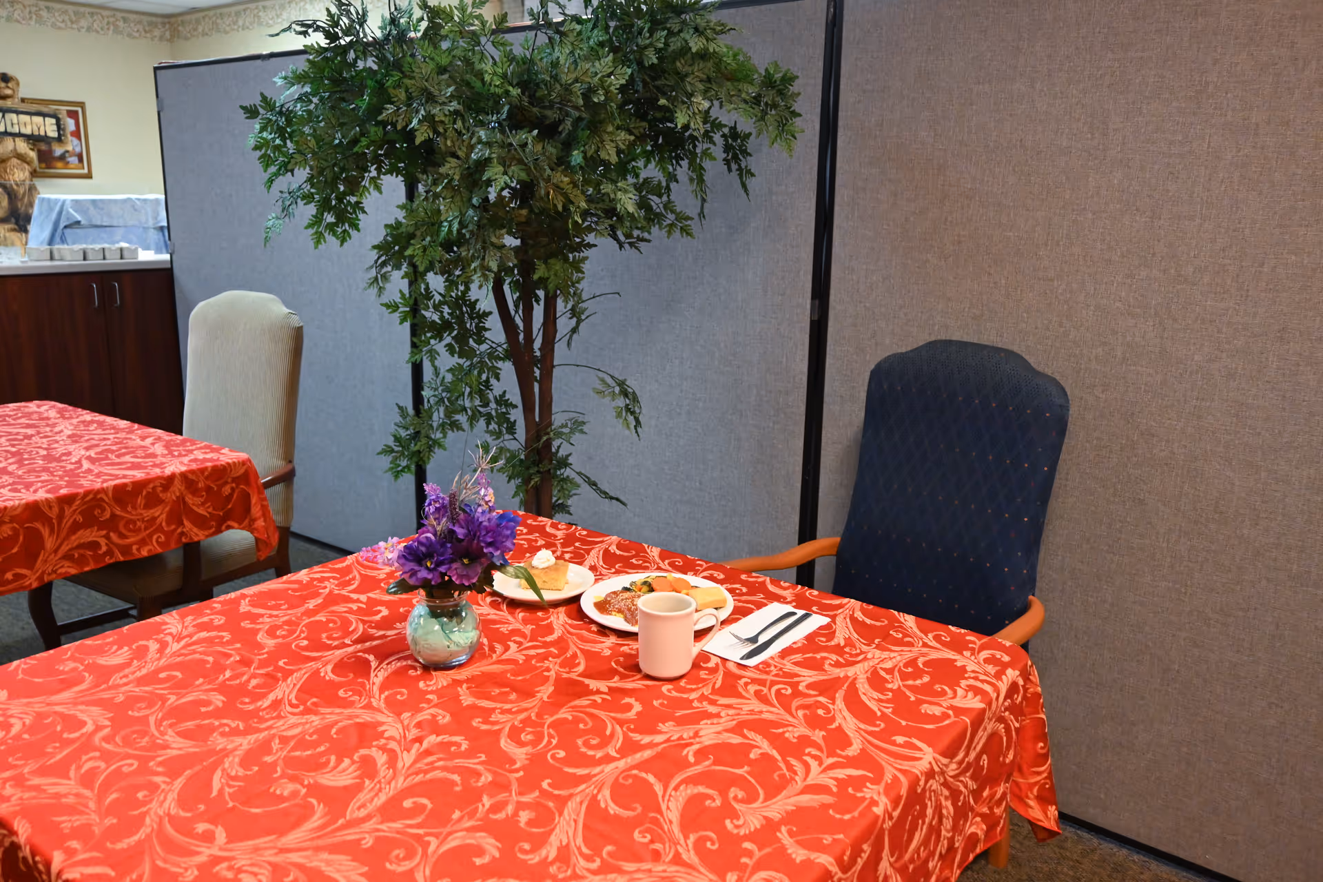 A dining table with a red patterned tablecloth, a small vase of purple flowers, a plate and cup, and chairs in a communal dining area.
