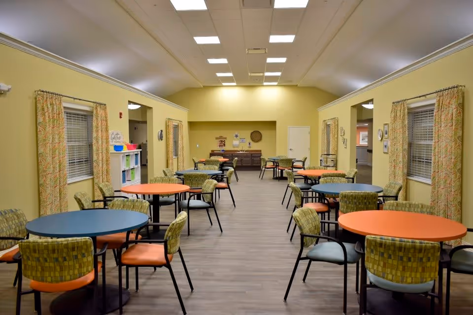 A spacious dining room with multiple round tables in blue and orange colors, each surrounded by chairs with patterned green and orange cushions. The walls are painted yellow and decorated with patterned curtains on the windows. The room has a wooden floor and bright ceiling lights.