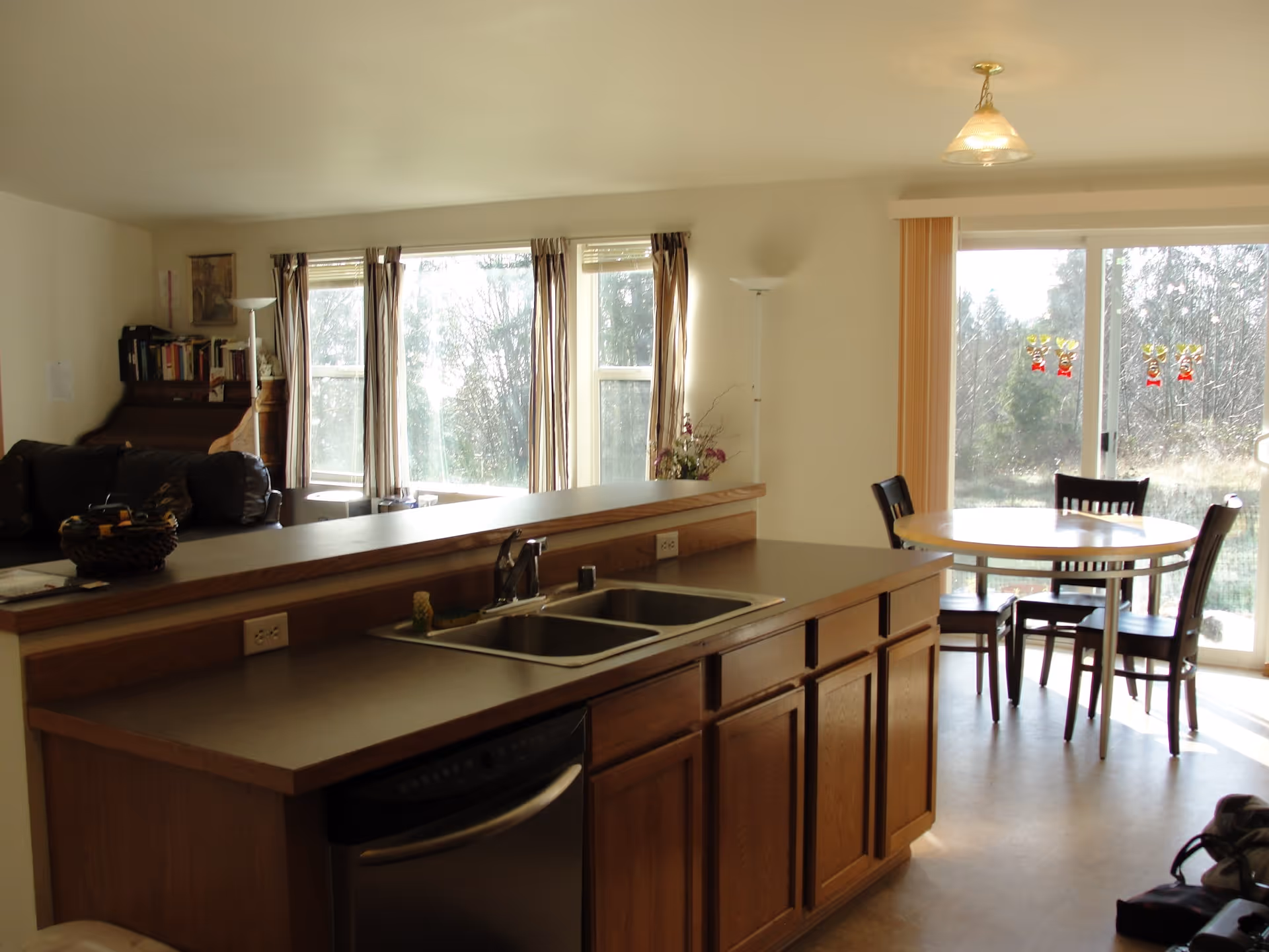 Interior view of a combined kitchen and dining area in a senior living facility. The kitchen features a countertop with a double sink, wooden cabinets, and a dishwasher. In the background, there is a round dining table with four chairs near a sliding glass door that leads outside. The living area with a couch and bookshelf is partially visible to the left, and large windows with curtains allow natural light to fill the space.