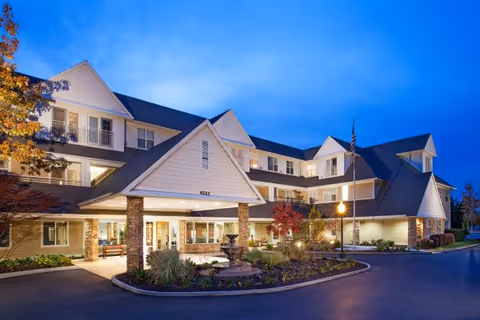 Exterior view of a large, well-lit senior living facility building at dusk with multiple windows, a covered entrance supported by stone pillars, landscaped garden with a fountain, and an American flag on a flagpole.