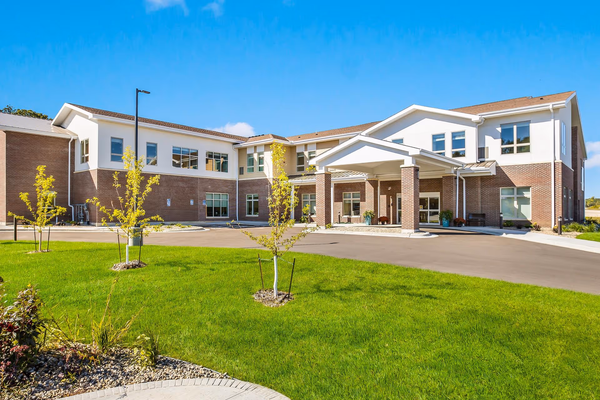 Exterior view of a two-story senior living facility building with a brick and white facade, a covered entrance supported by brick columns, young trees planted in a well-maintained green lawn, and a clear blue sky.