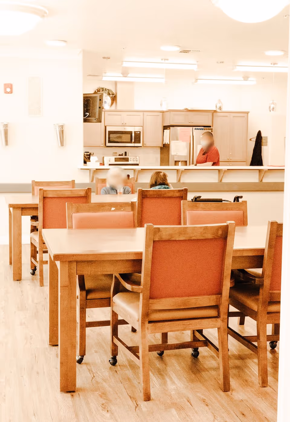 Interior view of a dining area in a senior living facility with wooden tables and chairs with orange cushions. In the background, there is a kitchen area with cabinets, a microwave, and a refrigerator. Two people are seated at a table, and one person is standing behind the counter.