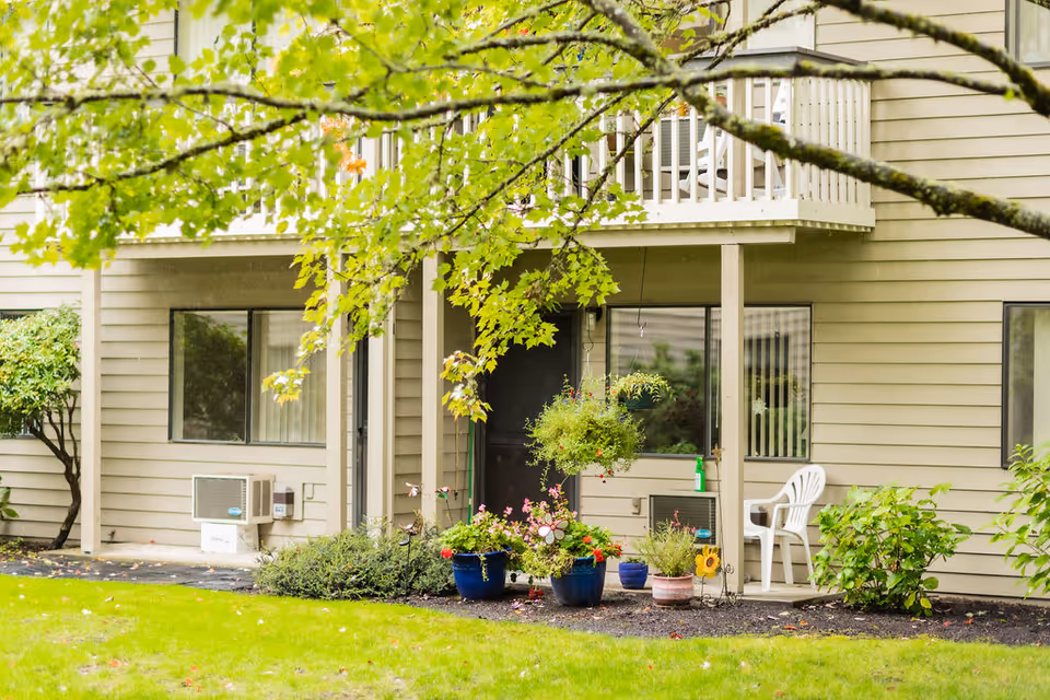 Exterior view of a beige two-story building with a small patio area on the ground floor featuring potted plants, a white plastic chair, and hanging flower baskets. Green grass and a tree branch with green leaves are visible in the foreground.