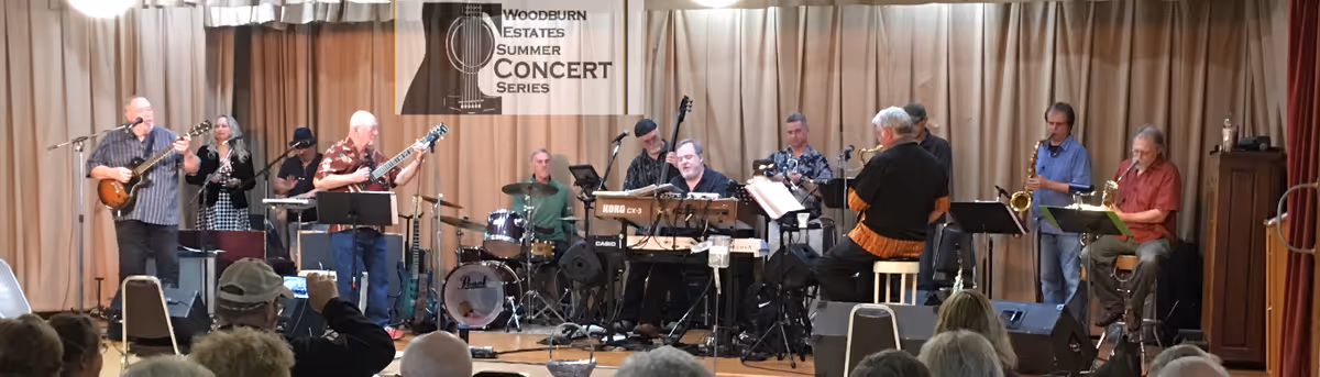 A musical band performing on stage with various instruments including guitars, drums, keyboards, saxophones, and a clarinet. The audience is seated in front of the stage. A banner behind the band reads 'Woodburn Estates Summer Concert Series'.