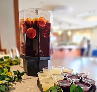 Large beverage dispenser filled with a red citrus punch and small plastic cups of red and yellow drinks on a decorated table in a dining area.