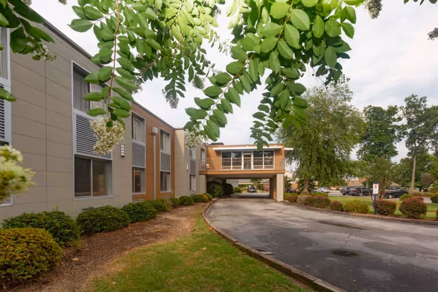 Exterior view of Millennium Post Acute Rehabilitation building with a driveway leading under a covered walkway. The building is surrounded by green bushes and trees, with a partly cloudy sky overhead.