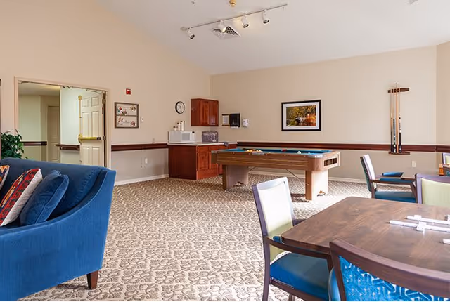 A spacious common room in an assisted living facility featuring a blue upholstered sofa, a pool table with cues on the wall, a wooden table with chairs, and a small kitchenette area with a microwave and coffee maker. The room has beige walls, patterned carpet, and a framed picture on the wall.