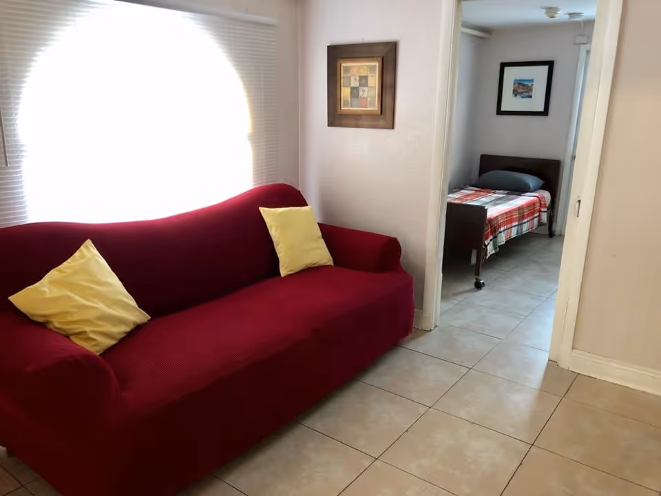 A small living area with a red sofa and yellow pillows in front of a bright arched window, opening into a bedroom with a single bed.