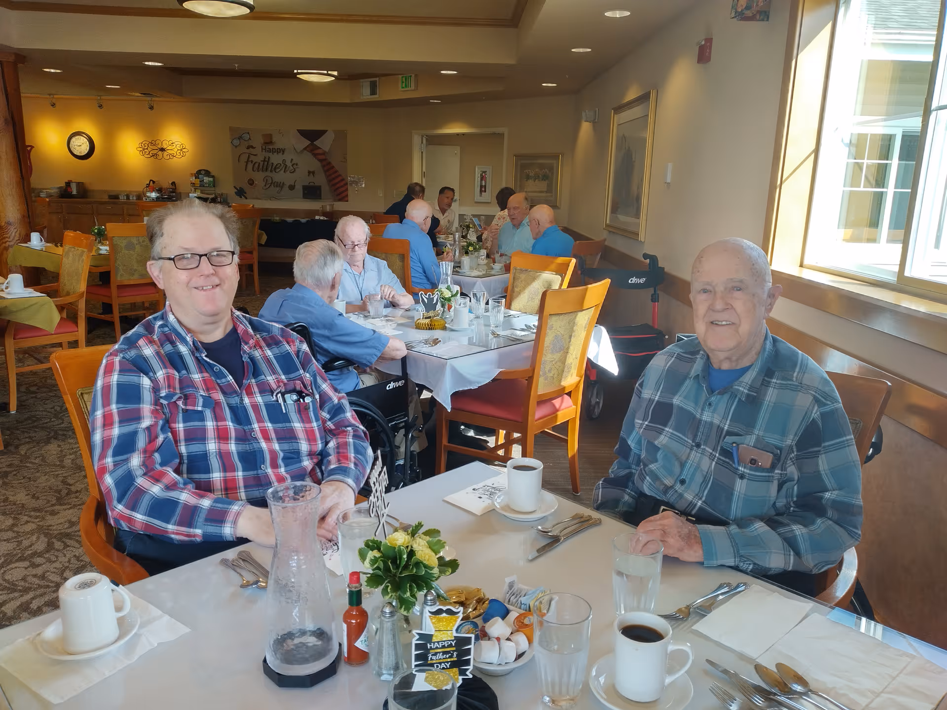 Elderly residents sit and smile around a table set for a meal in a bright communal dining room.