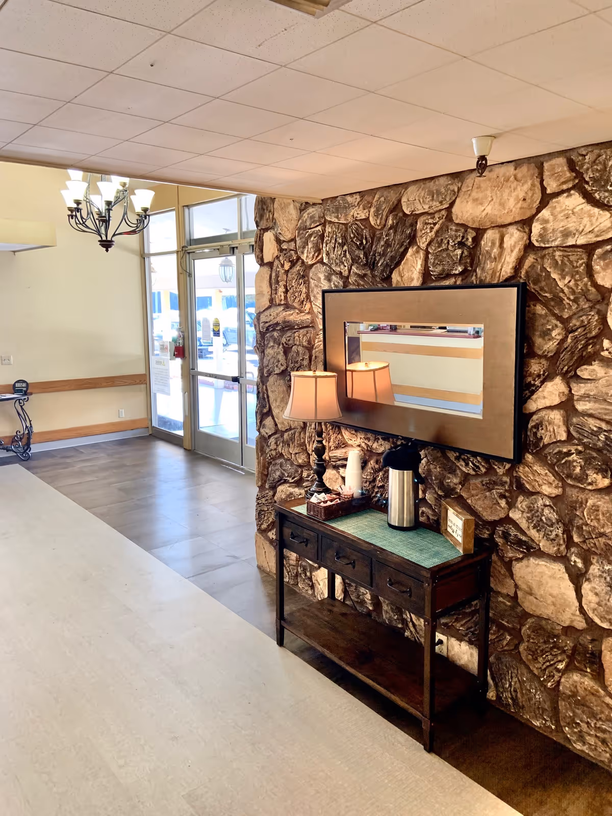 Interior view of a facility entrance area with a stone accent wall. A wooden console table with three drawers holds a lamp, a coffee thermos, cups, and a small sign. Above the table is a rectangular mirror. Glass double doors lead outside, and a chandelier hangs from the ceiling.