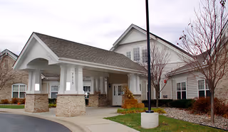 Exterior view of a senior living facility building with a covered entrance supported by white columns and brick bases. The building has white siding, multiple windows, and a gabled roof. There are leafless trees and landscaping with rocks and grass around the entrance area.