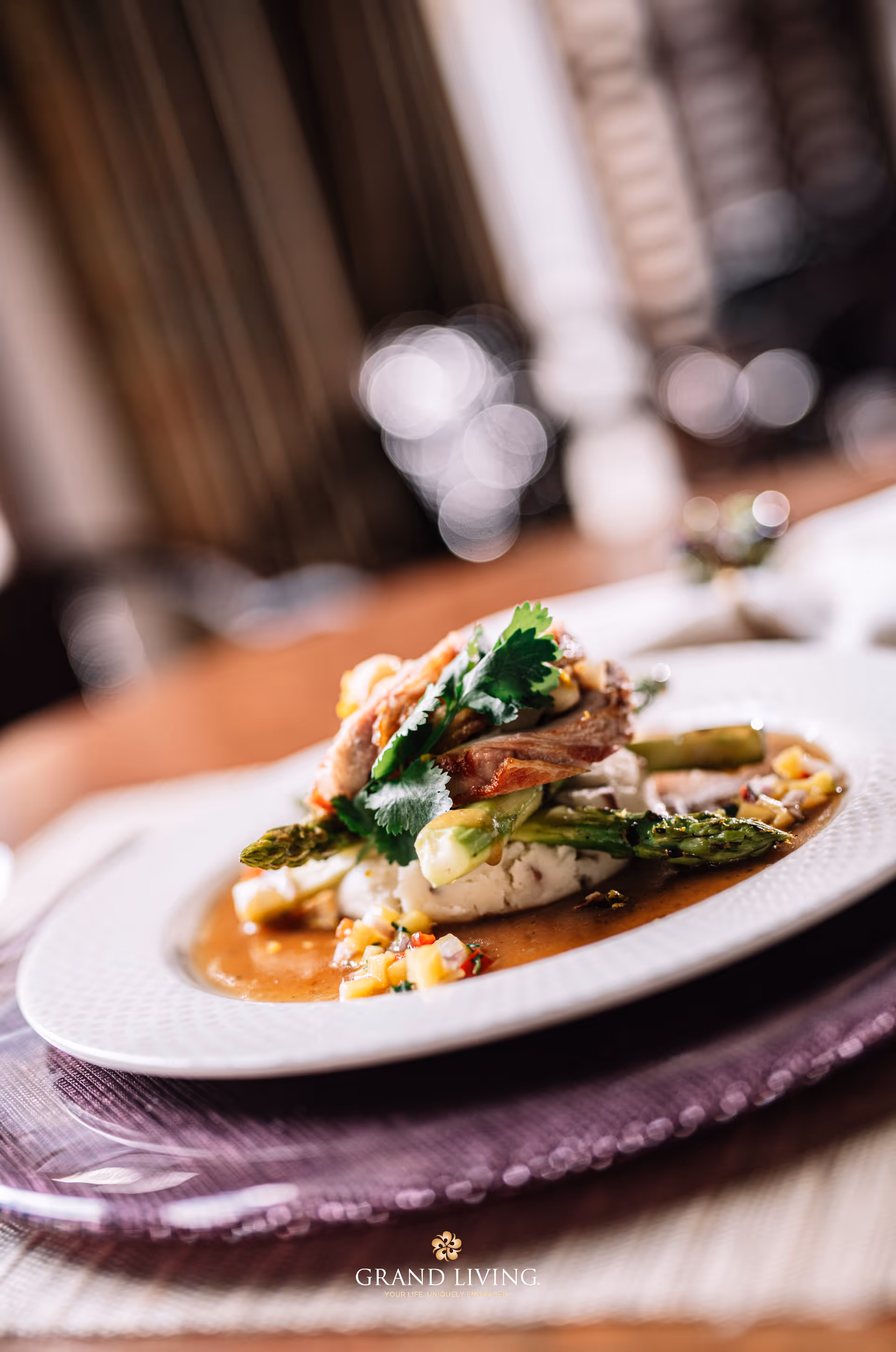 Close-up of a gourmet dish featuring grilled asparagus, mashed potatoes, and a piece of meat garnished with fresh herbs, served on a white plate with sauce and diced vegetables, placed on a purple placemat on a wooden table.