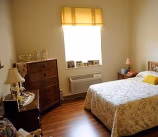 A cozy bedroom with a bed covered in a floral quilt, a wooden dresser, a nightstand with a lamp, and a window with a yellow valance letting in natural light.