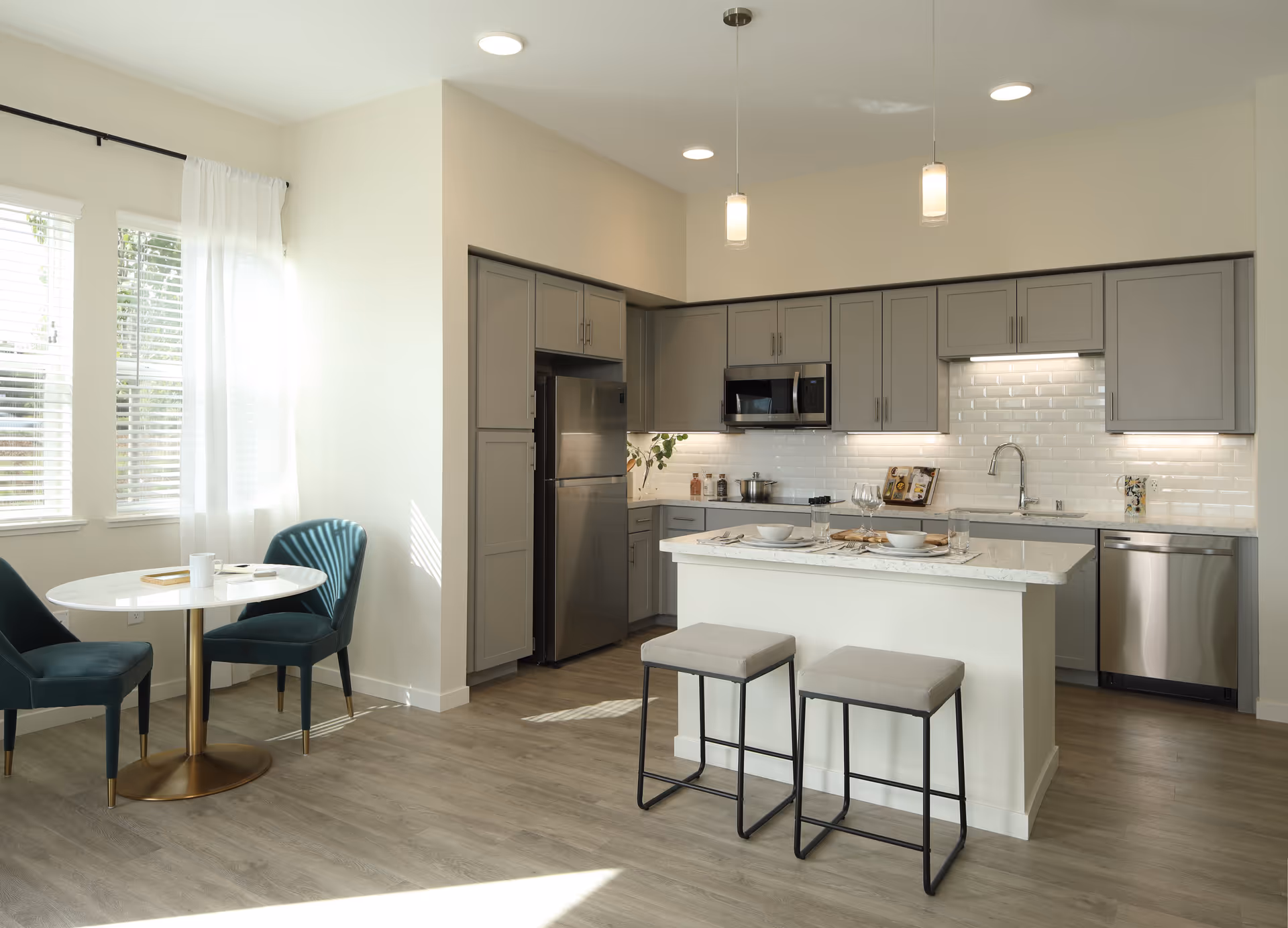 Bright modern kitchen with gray cabinets, stainless steel appliances including refrigerator, microwave, and dishwasher. A white island with marble countertop has two stools. To the left, there is a small round dining table with two teal upholstered chairs near windows with white curtains.