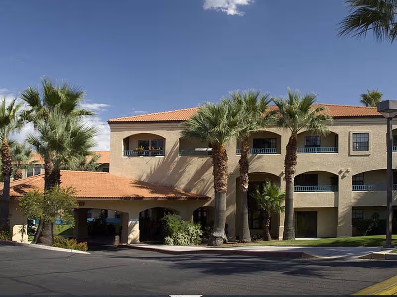Exterior view of a two-story senior living facility building with beige walls and a red-tiled roof, surrounded by palm trees and landscaping under a clear blue sky.