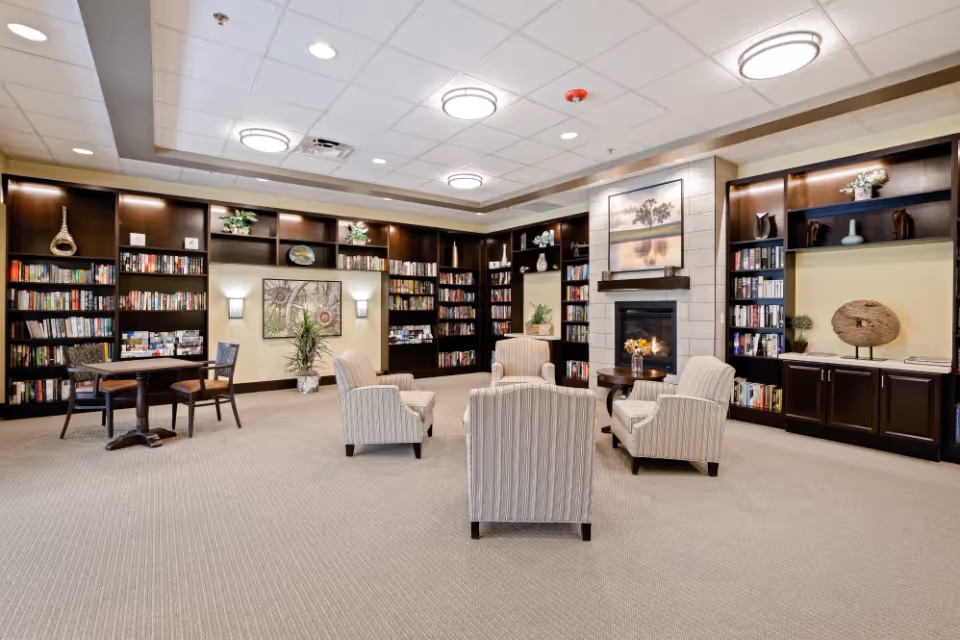 A spacious and well-lit common area with beige carpet and a white ceiling with recessed lighting. The room features dark wood built-in bookshelves filled with books and decorative items along the walls. In the center, there are four striped upholstered armchairs arranged around a small round wooden table with a flower arrangement. A fireplace with a large framed artwork above it is visible on the right side of the room. A small round table with two chairs is positioned near the bookshelves on the left side. The overall atmosphere is cozy and inviting.
