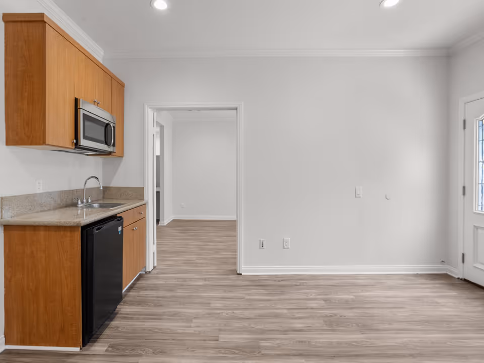 Interior view of a small kitchen area with wooden cabinets, a countertop with a sink, a microwave mounted above, and a mini refrigerator below. The kitchen opens into an empty room with light wood flooring and white walls. A door with glass panels is visible on the right side.