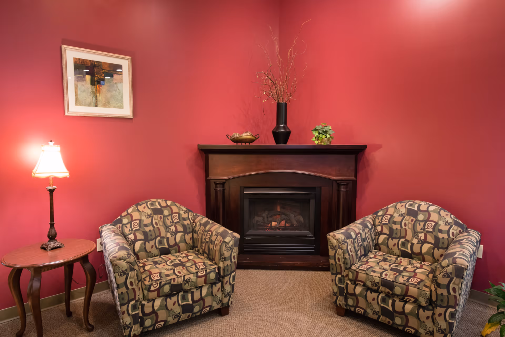 A cozy sitting area with two patterned armchairs facing a dark wooden electric fireplace. The walls are painted a deep red, and there is a small wooden side table with a lamp on the left. Above the fireplace are decorative items including a black vase with branches and a small green plant. A framed abstract painting hangs on the wall to the left.