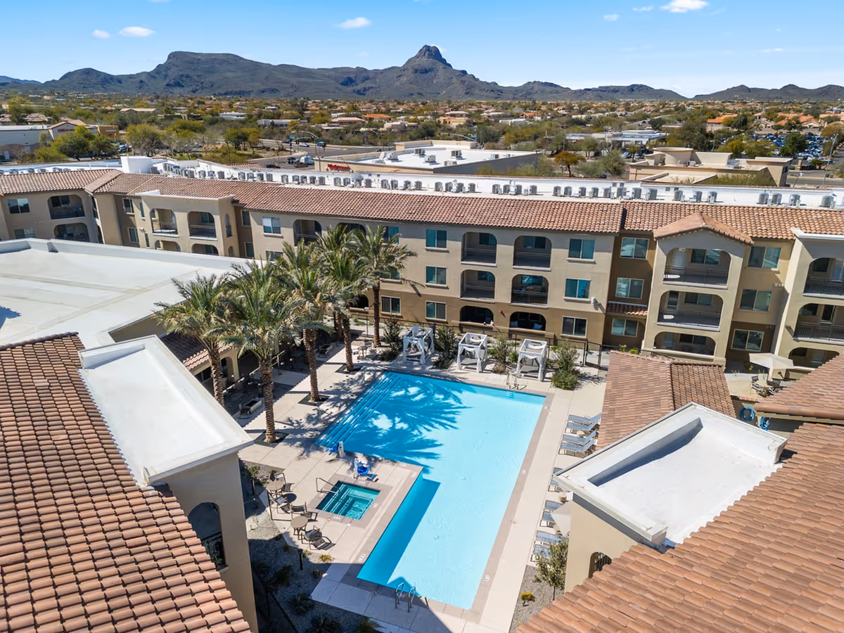 Aerial view of a senior living facility courtyard with a large swimming pool, a smaller hot tub, palm trees, lounge chairs, and cabanas. The surrounding building has multiple floors with balconies and a tiled roof. Mountains and a suburban area are visible in the background under a clear blue sky.