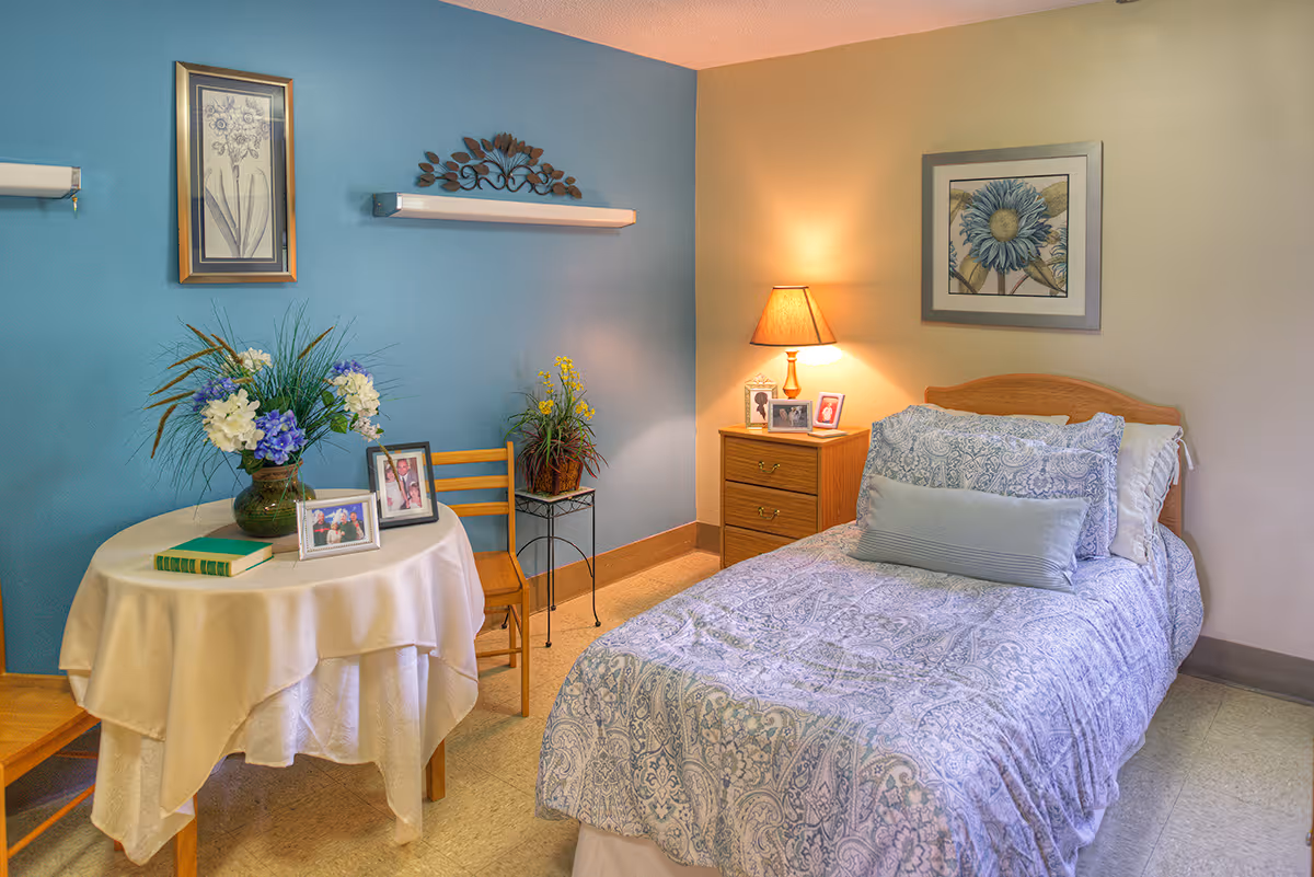 A neatly decorated single-occupancy bedroom with a bed, nightstand and lamp, and a round table with flowers and framed photos against a teal accent wall.