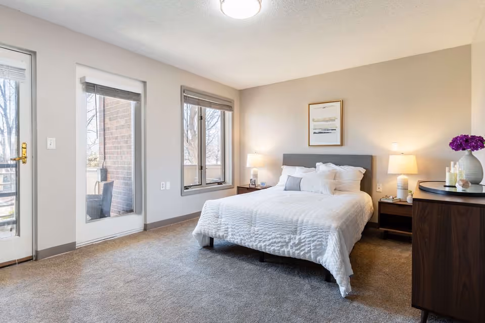 A bright and clean bedroom with a large bed dressed in white bedding, two bedside tables with lamps, a framed abstract artwork above the bed, a window and glass door leading to an outdoor balcony, and a wooden dresser with a vase of purple flowers and decorative items.