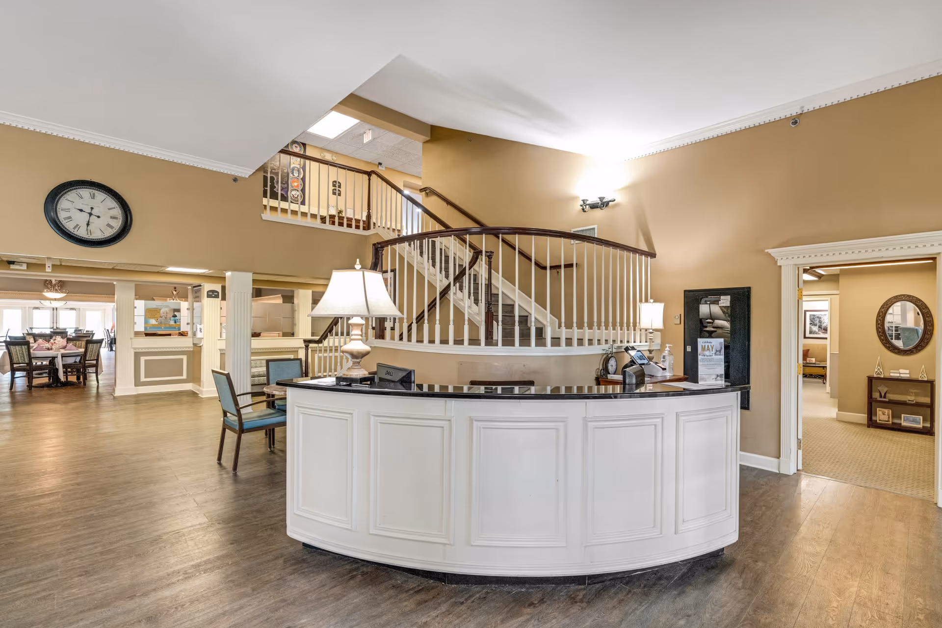Interior view of a senior living facility reception area with a curved white front desk, two table lamps, and a staircase with white railings leading to an upper floor. To the left, there is a dining area with tables and chairs, and a large wall clock is mounted above the entrance to the dining space. The walls are painted beige, and the floor has a wood finish.
