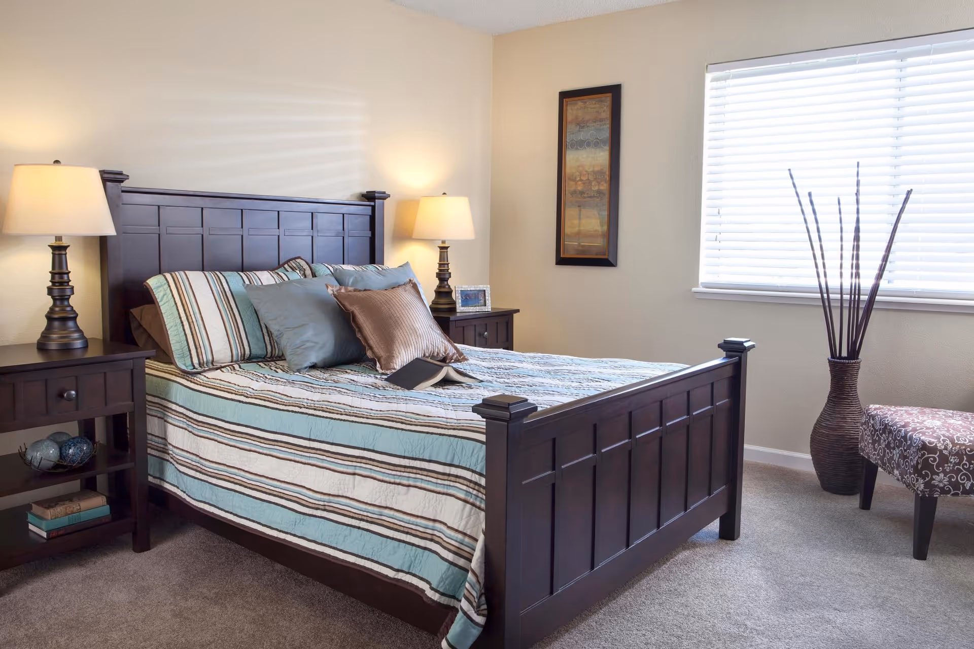 Well-lit bedroom with a dark wood bed and matching nightstands, striped bedding, two lamps, a window with blinds, and decorative accents.