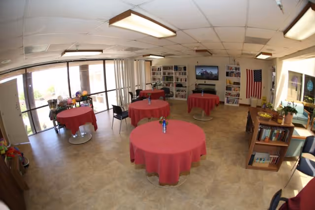A bright common room with round tables covered in red tablecloths, each with a small vase of flowers. The room has large windows letting in natural light, bookshelves filled with books, a television mounted on the wall, an American flag, and some chairs arranged around the tables. There are also some plants and a small bookshelf near the seating area.