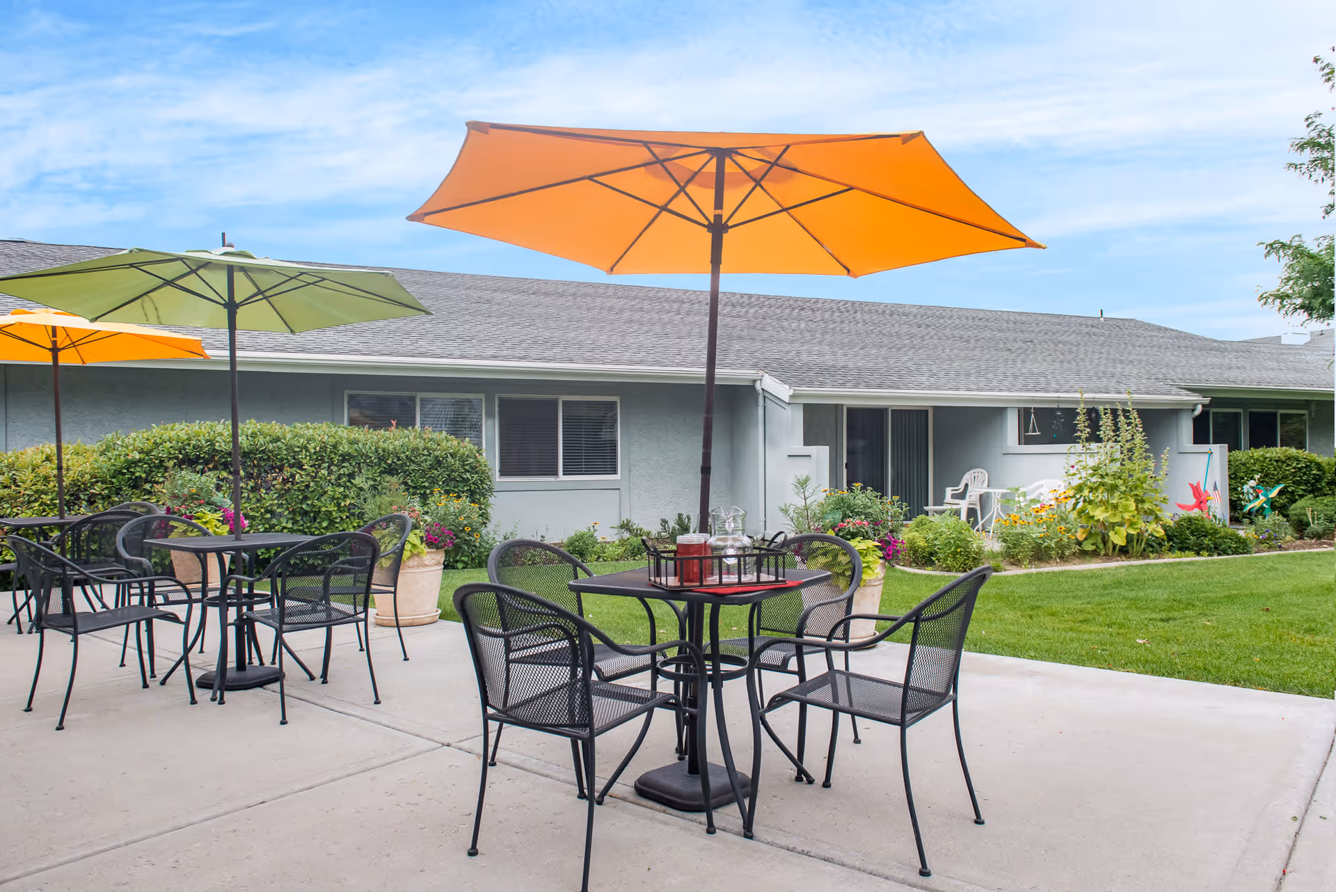 Outdoor patio area at Highland Cove Retirement with black metal tables and chairs under colorful umbrellas, surrounded by green grass, bushes, and flowers in front of a single-story building.