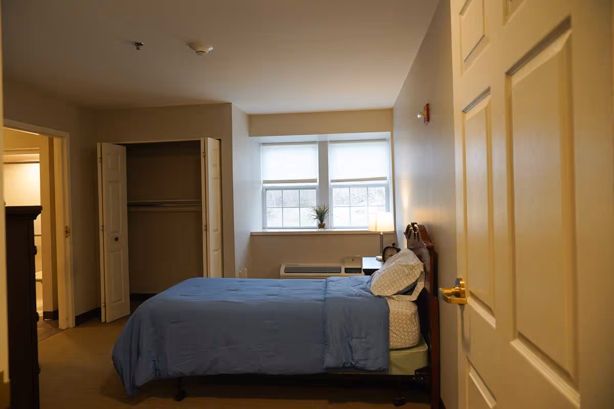 A bedroom with a bed covered in a blue comforter and white pillows. There is a window with two blinds partially drawn, a small plant on the windowsill, and a bedside table with a lamp and an alarm clock. An open closet with bi-fold doors is visible, as well as a doorway leading to a bathroom.