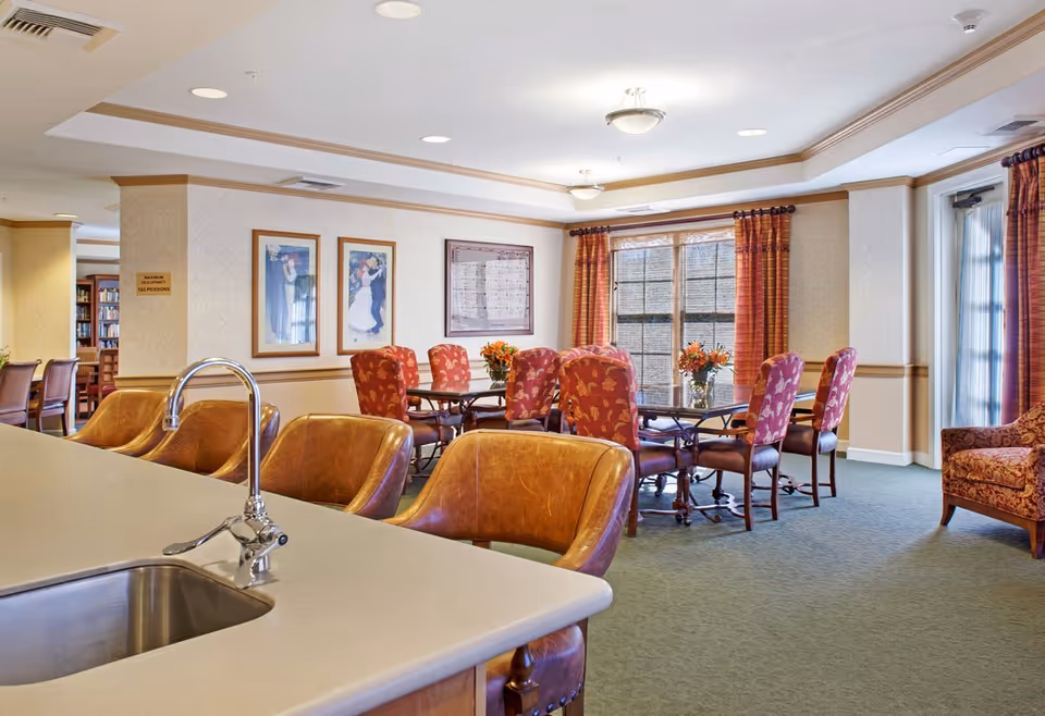 Communal dining and lounge area with a countertop and sink in the foreground, leather bar stools, and a round table with upholstered chairs by windows.