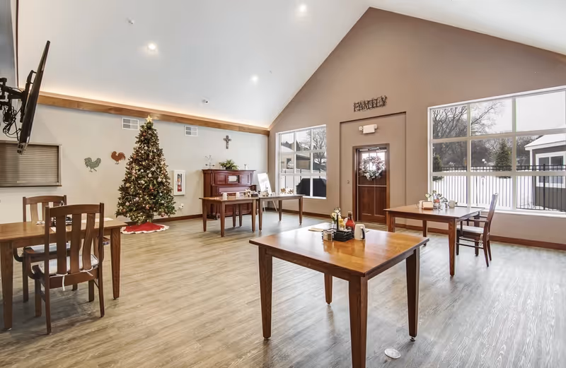 A bright and spacious dining area with wooden tables and chairs, a decorated Christmas tree, a piano, and large windows letting in natural light. The room has a high vaulted ceiling and a door with a wreath, with the word 'FAMILY' displayed above it.