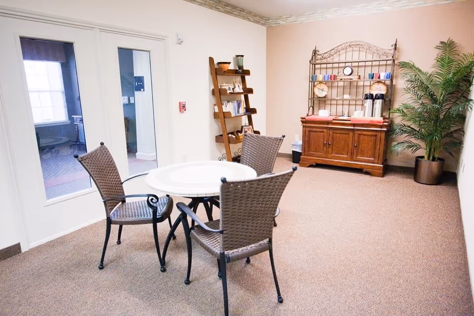 A small seating area with a round table and three wicker chairs on a carpeted floor. Behind the seating area is a wooden cabinet with a metal rack holding cups, plates, and coffee dispensers. There is a tall potted plant to the right and a wooden shelf with books and decorative items against the wall. Double glass doors are visible on the left side of the room.