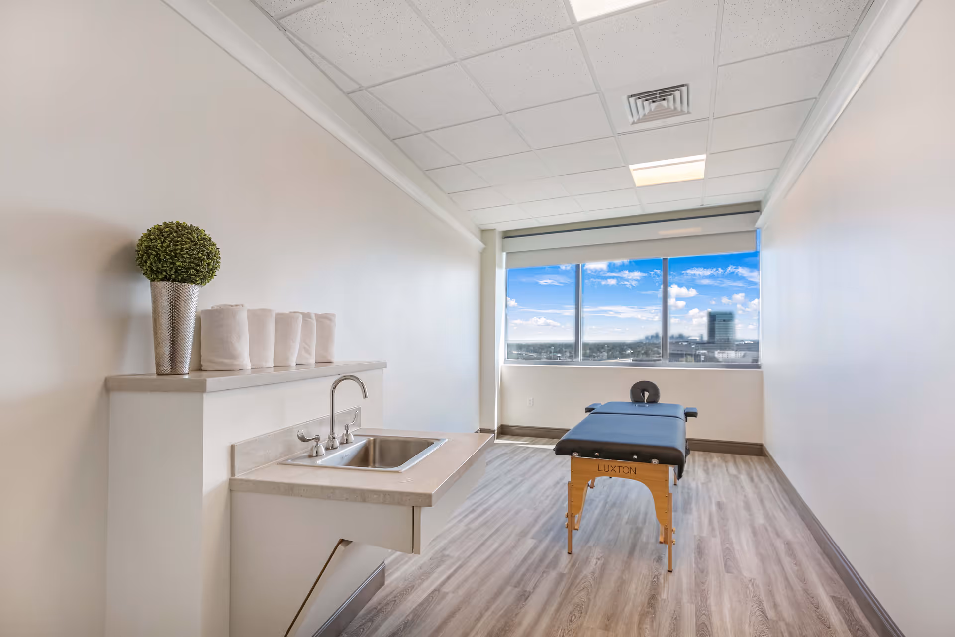A bright room with a large window showing a cityscape view. Inside the room, there is a massage table with a face cradle positioned near the window. On the left side, there is a countertop with a small sink, a potted plant, and several rolled white towels. The room has light-colored walls and wood-patterned flooring.