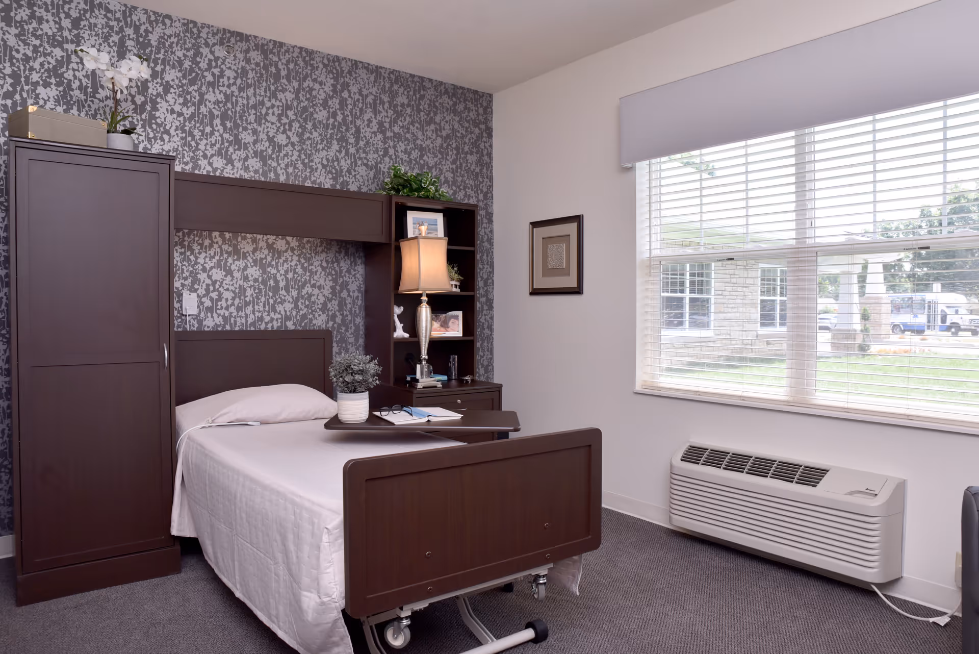 A neatly arranged bedroom in a senior living facility with a single bed featuring a white bedspread, a dark wooden headboard and footboard, and an overbed table with a potted plant and reading materials. Behind the bed is a dark wooden storage unit with shelves holding a lamp, framed photos, and decorative items. The wall behind the bed has a patterned wallpaper, and there is a large window with blinds letting in natural light. Below the window is a white air conditioning unit.