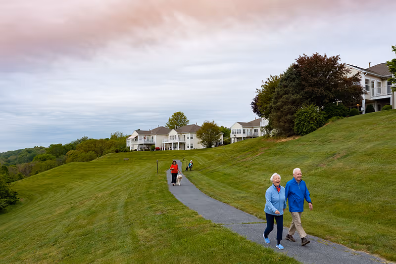 A paved walking path through a grassy hillside with several senior individuals walking and enjoying the outdoors near residential buildings under a cloudy sky.