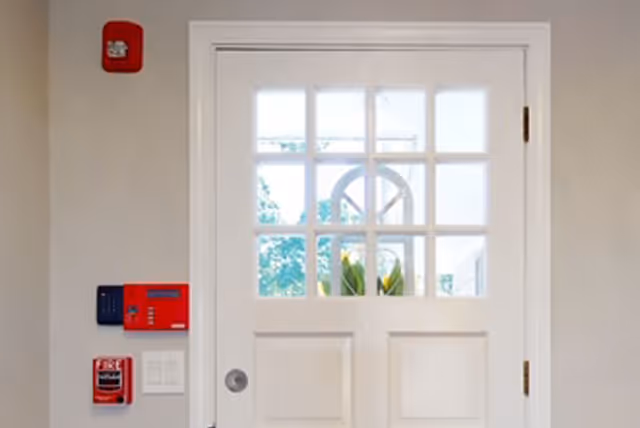 White door with nine glass panes at the top, showing a glimpse of greenery outside. To the left of the door are a red fire alarm, a red fire alarm pull station, and a black keypad mounted on a beige wall.