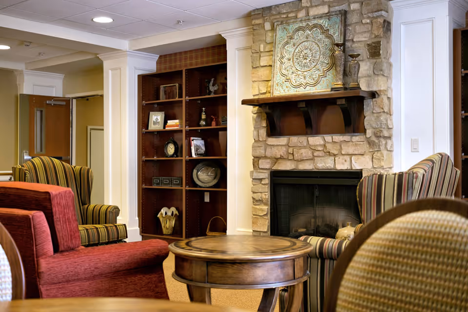 Cozy living room area with a stone fireplace, wooden mantel, and decorative artwork above it. The room features comfortable upholstered chairs in striped and solid patterns, a round wooden coffee table, and built-in wooden shelves with various decorative items and books.