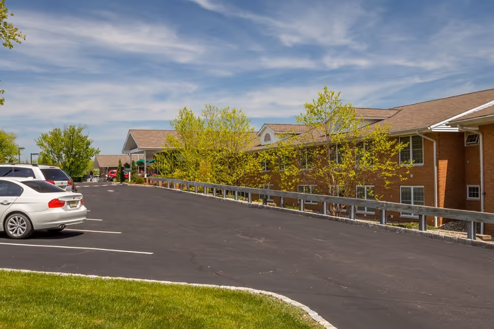 Parking lot with several parked cars next to a two-story brick building with multiple windows and a sloped roof. Trees with green leaves are visible along the building, and the sky is partly cloudy.