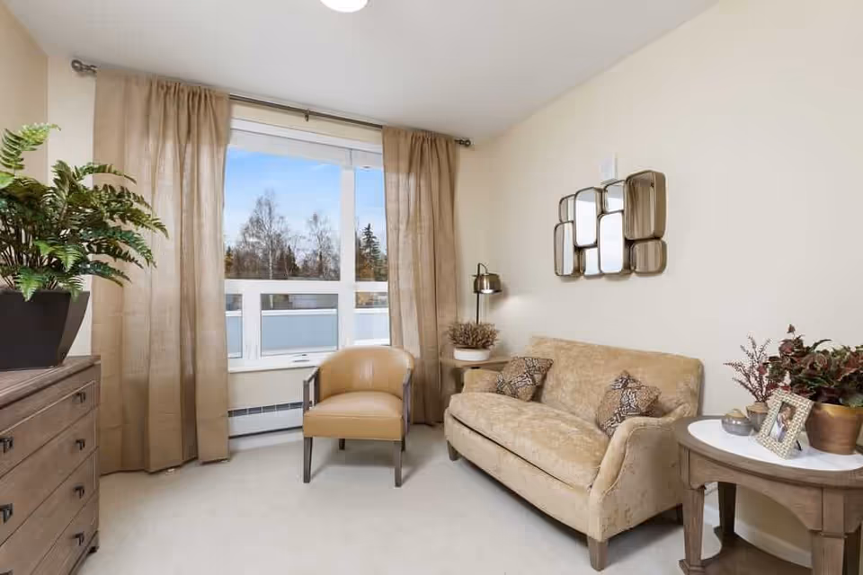 Sunlit sitting room with a sofa, armchair, side tables, plants, and a window with curtains.