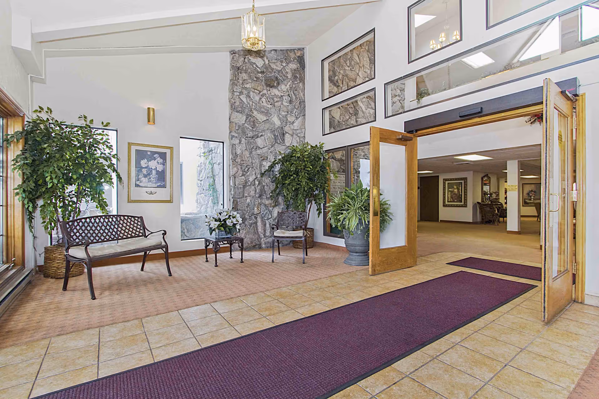 Entrance area of a retirement community featuring a tiled floor with a purple runner rug, wooden double doors open to an interior space with carpeted floors. The area has a stone accent wall, framed floral artwork, several potted plants, a metal bench with a cushion, and a metal chair with a cushion. Large windows and mirrors reflect the interior light.