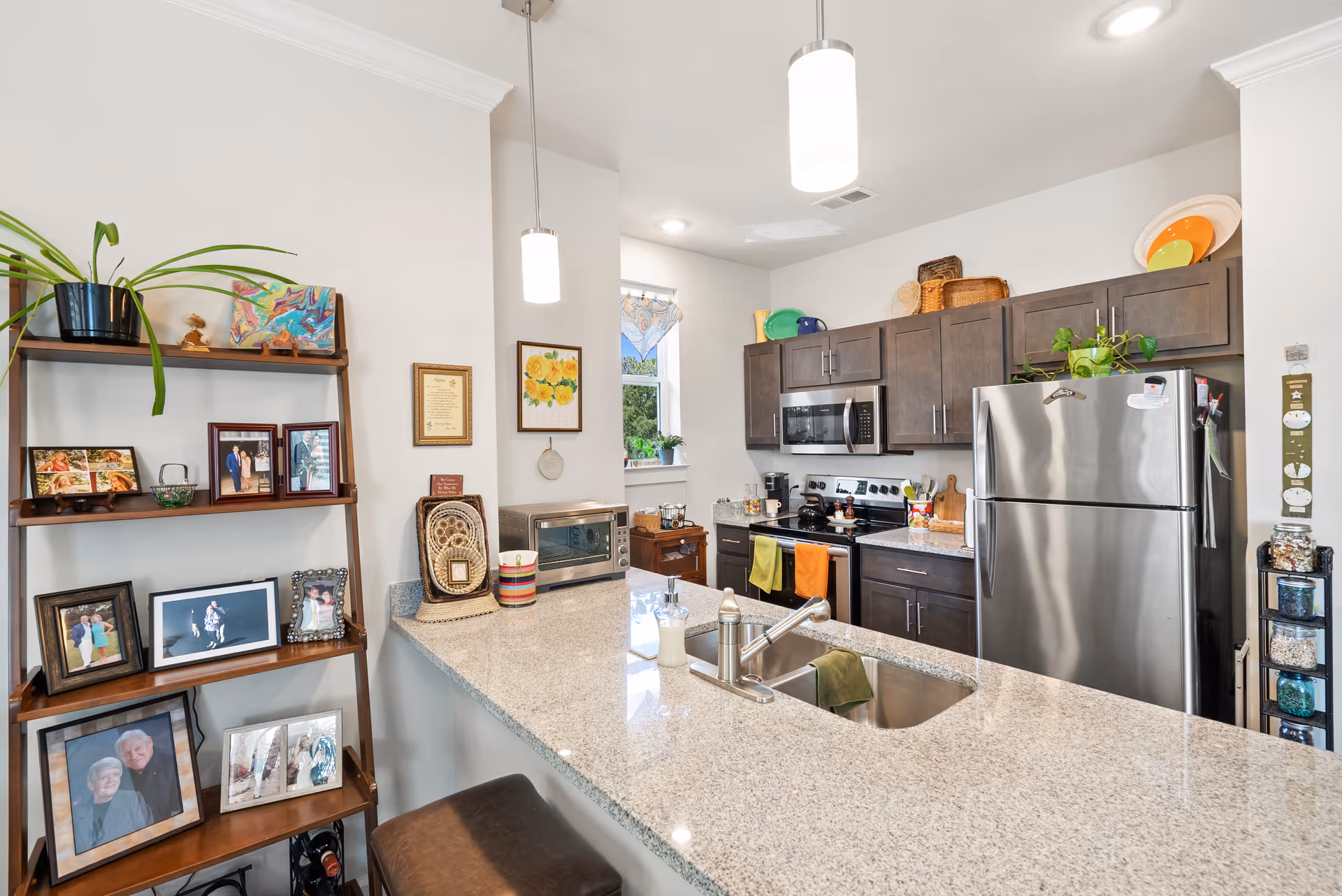 Modern kitchen with granite countertop island featuring a sink and faucet. Dark wood cabinets, stainless steel refrigerator, microwave, and stove with colorful towels hanging on the oven handle. A wooden shelf on the left holds framed photos and a potted plant. The kitchen is well-lit with two pendant lights and a small window above the counter.
