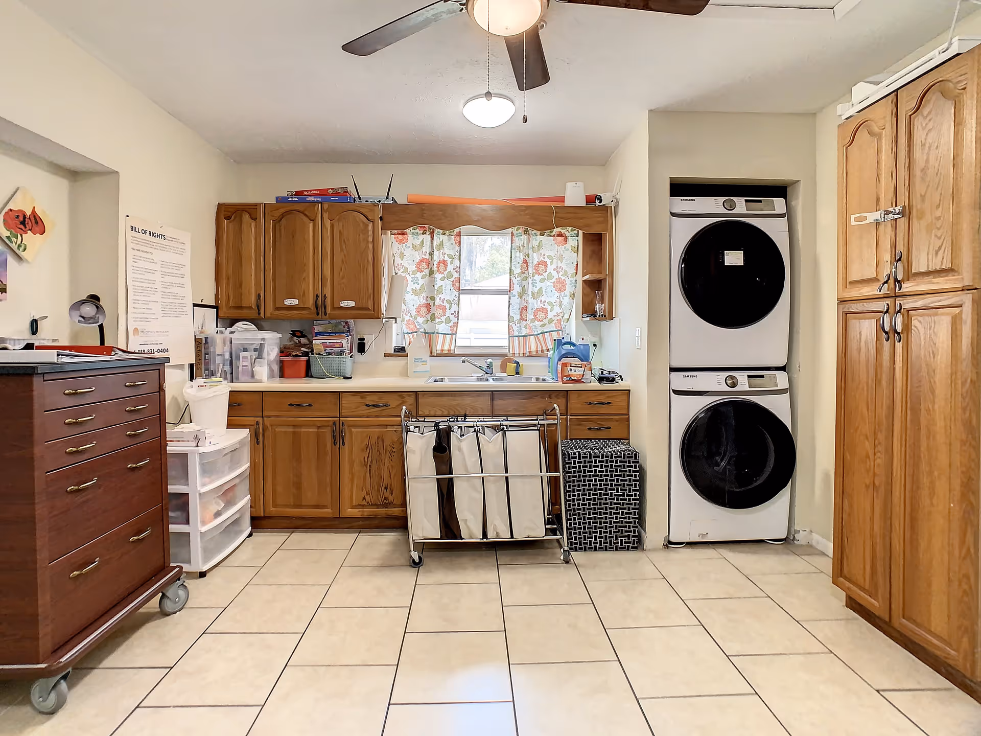 Laundry room with tiled floor, wooden cabinets, a stacked washer and dryer, a laundry sorter cart, and a window with floral curtains above a sink.