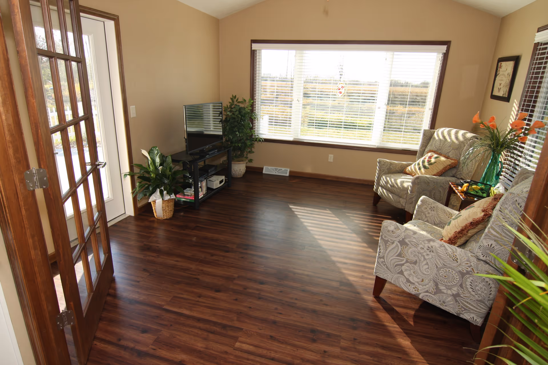 A bright living room with wooden flooring, two patterned armchairs with cushions, a small side table with a vase of orange flowers, a flat-screen TV on a black stand, and two potted plants near a large window with white blinds letting in sunlight.