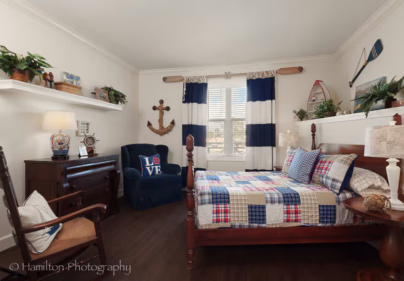 A cozy bedroom with a wooden bed featuring a patchwork quilt in red, blue, and beige tones. The room has dark wood flooring, a navy blue armchair with a pillow that says 'LOVE', and nautical-themed decor including an anchor on the wall and oars above the window. There are white shelves with plants and decorative items, a wooden dresser with a lamp, and a window with blue and white striped curtains letting in natural light.