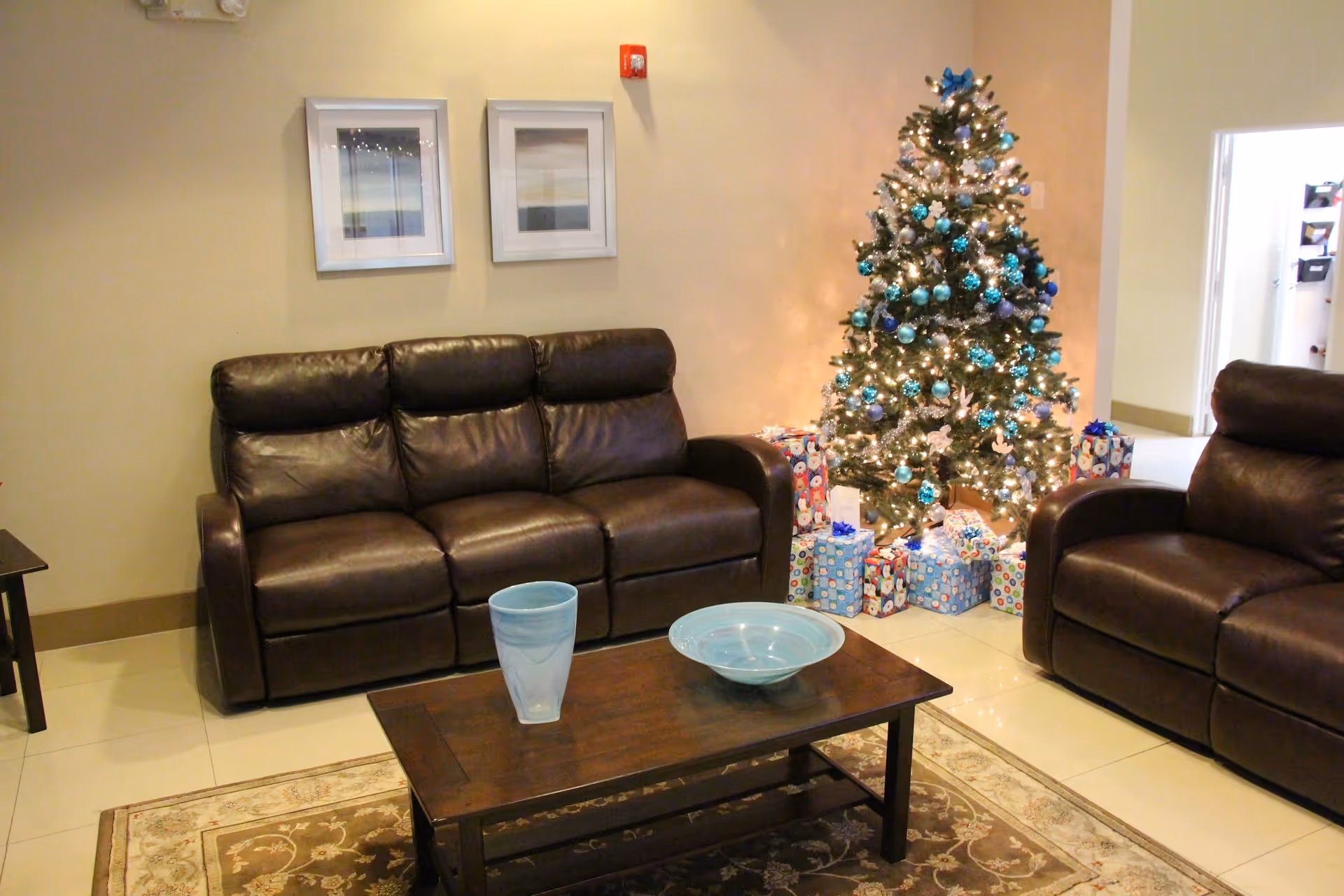 A cozy living room area with two dark brown leather sofas facing a wooden coffee table. On the table, there is a blue glass and a blue bowl. In the background, a decorated Christmas tree with blue and silver ornaments and lights is surrounded by wrapped presents. The walls are light-colored with two framed pictures hanging above one sofa.