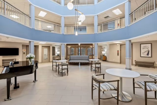Spacious and bright common area inside a senior living facility with a grand piano on the left, several round tables with chairs, a gray sofa against a decorative wooden screen, and a three-story atrium with railings and hanging globe lights.