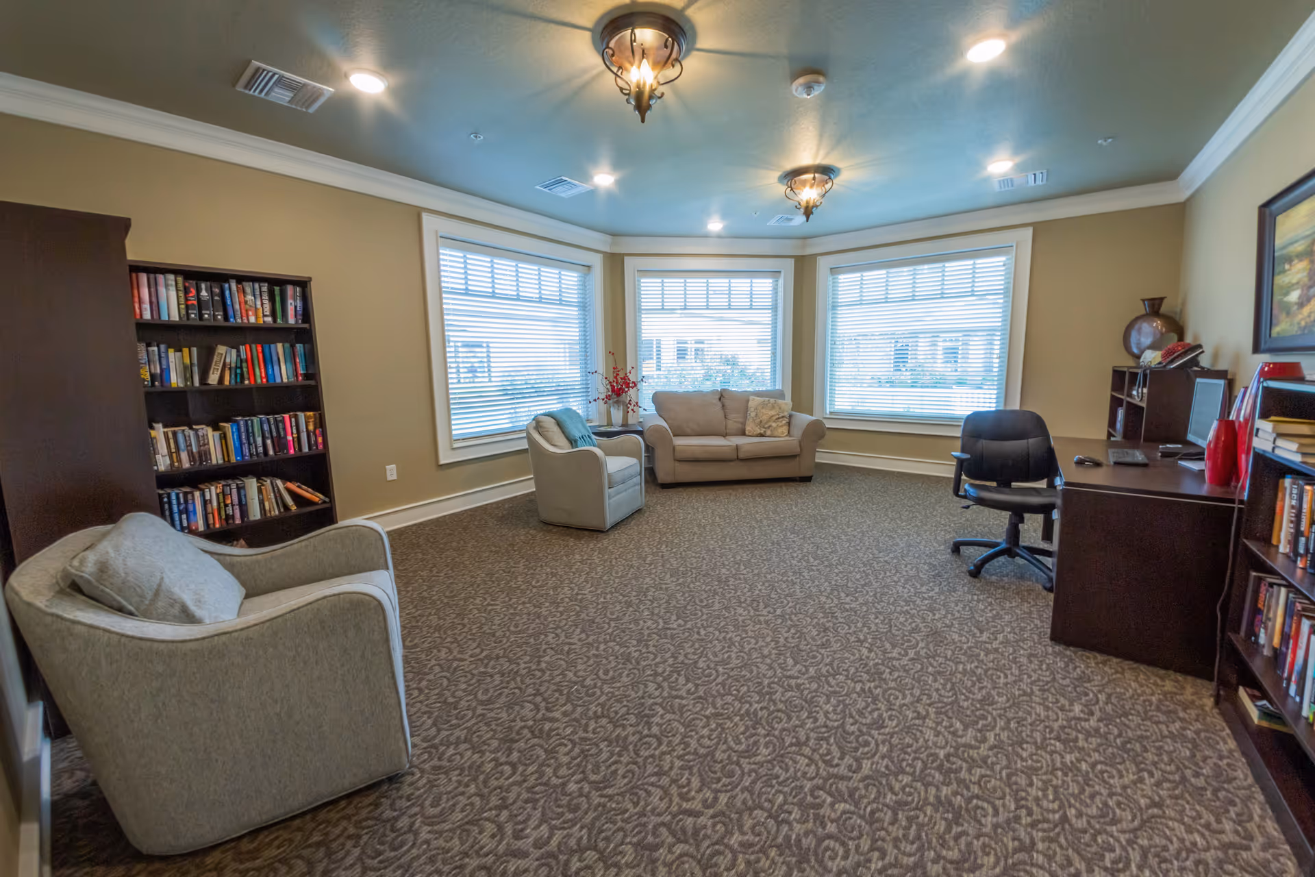 A cozy sitting room with beige walls and carpeted floor featuring a beige loveseat, a gray armchair, a dark wood desk with a black office chair, and two dark wood bookshelves filled with books. Three large windows with white blinds allow natural light into the room. The ceiling has decorative light fixtures and recessed lighting.